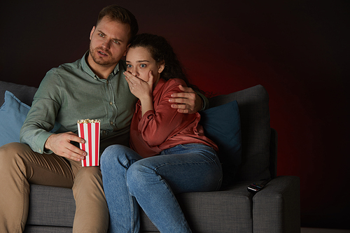 Portrait of young couple watching horror movie in dark room, focus on scared young woman, copy space
