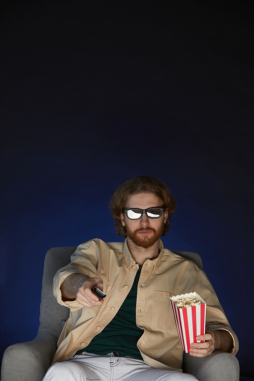 Vertical portrait of modern bearded man watching movie and wearing stereo glasses while sitting on couch in dark room, copy space