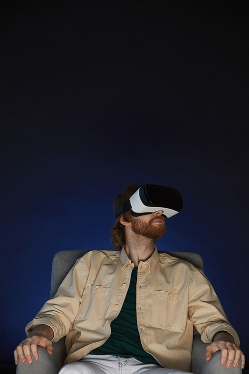 Vertical portrait of modern bearded man wearing virtual reality gear while enjoying immersive videogame or movie in dark, copy space