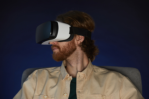 Side view portrait of modern bearded man wearing virtual reality gear while enjoying immersive videogame or movie in dark