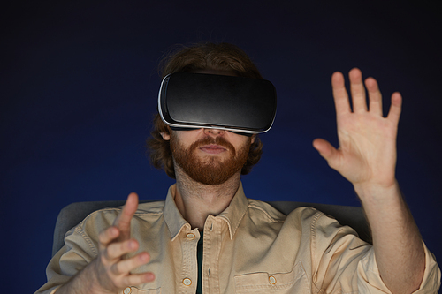 Head and shoulders portrait of modern bearded man wearing virtual reality gear while enjoying immersive videogame or movie in dark
