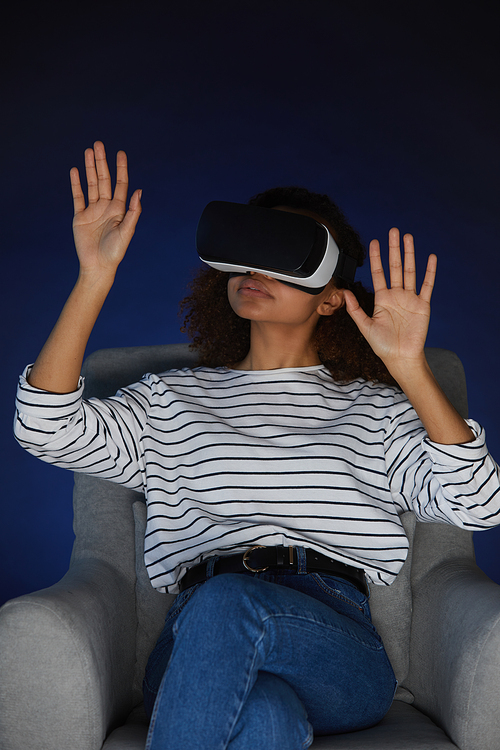 Vertical portrait of mixed-race young woman wearing virtual reality gear while enjoying immersive videogame or movie in dark, copy space