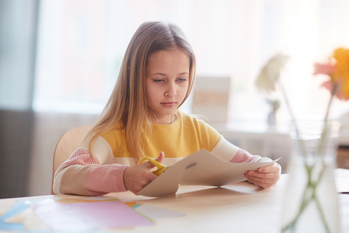 Portrait of cute girl cutting handmade holiday card for Mothers day or Valentines day while sitting at table with flowers, copy space