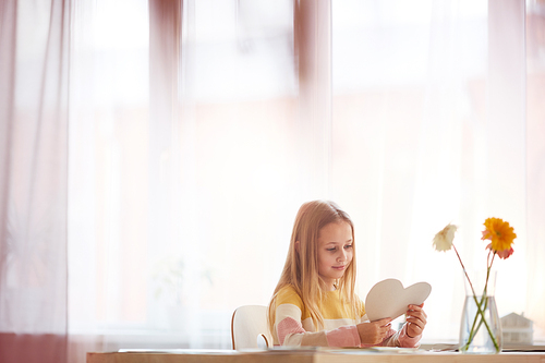 Warm-tones portrait of cute girl holding heart-shaped card while sitting at table with flowers on Valentines day, copy space