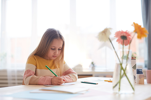 Portrait of cute little girl drawing pictures or doing homework while sitting at table in home interior, copy space