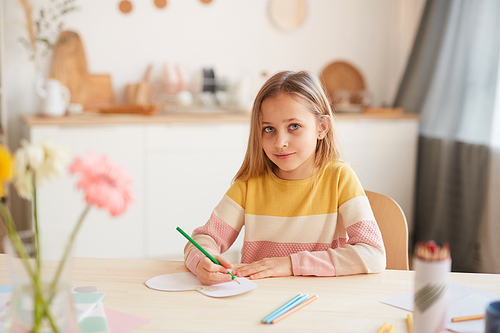 Warm toned portrait of cute little girl  and smiling while drawing pictures or doing homework while sitting at table in home interior, copy space