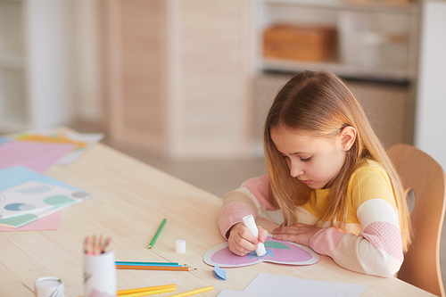 High angle portrait of cute little girl making handmade card for Mothers day while sitting at table in cozy home interior, copy space