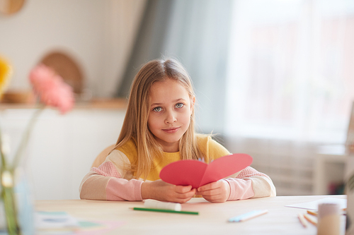 Portrait of cute little girl holding heart-shaped card and  while sitting at table in cozy home interior, copy space