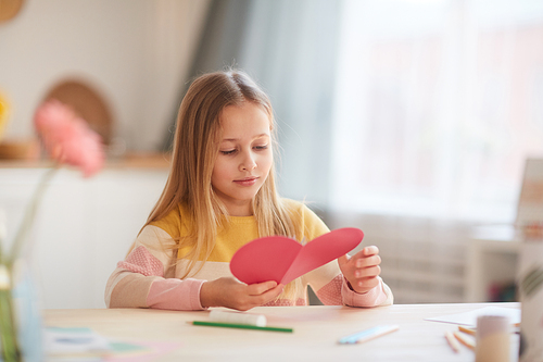 Portrait of cute little girl holding heart-shaped card and smiling while sitting at table in cozy home interior, copy space