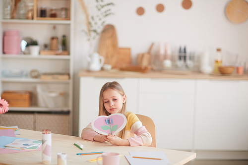 Wide angle portrait of cute little girl holding heart-shaped card while sitting at table in cozy home interior, copy space