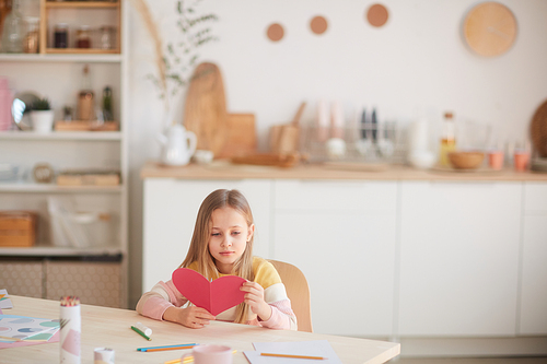 Wide angle portrait of cute blonde girl holding heart-shaped card while sitting at table in cozy home interior, copy space