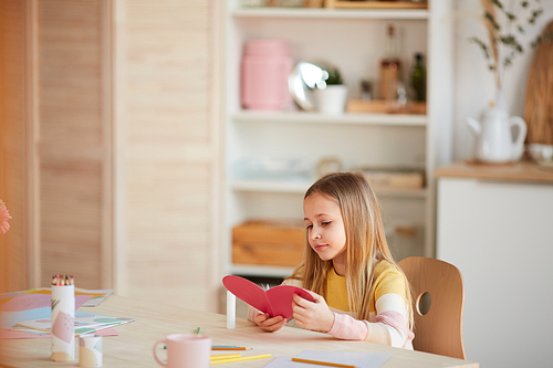 Warm-toned portrait of cute girl making heart-shaped card and smiling while sitting at table in cozy home interior, copy space