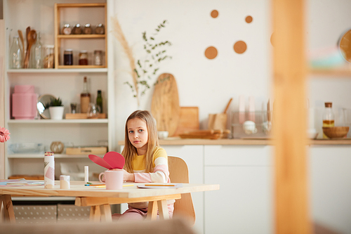 Wide angle portrait of cute girl holding heart-shaped card and  while sitting at table in cozy home interior, copy space