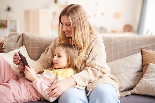 Warm-toned portrait of modern mother playing with cute girl while enjoying time together on couch and using digital tablet, copy space