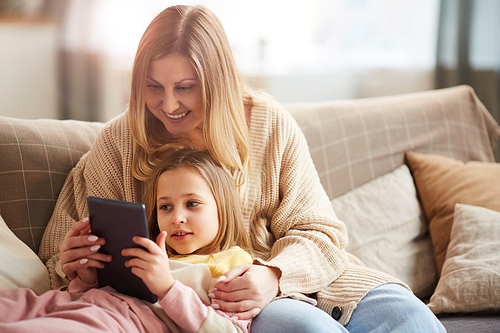 Warm-toned portrait of happy mature mother playing with cute girl while enjoying time together on couch and using digital tablet, copy space