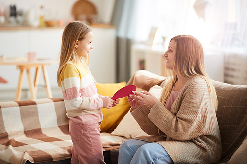 Side view portrait of cute little girl giving handmade card to mom on Mothers day in cozy home interior, copy space