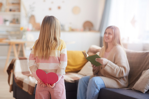 Back view portrait of cute girl holding handmade card for mom while surprising her on Mothers day in cozy home interior, copy space