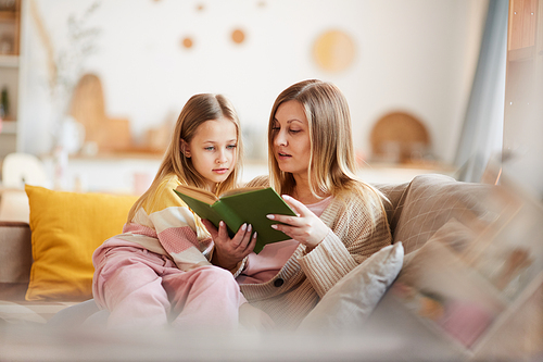 Warm-toned portrait of mature mother reading book to little girl while sitting on sofa in cozy home interior, copy space