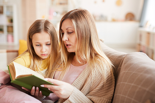 Warm-toned portrait of mature mother reading book to little girl while sitting on sofa in cozy home interior