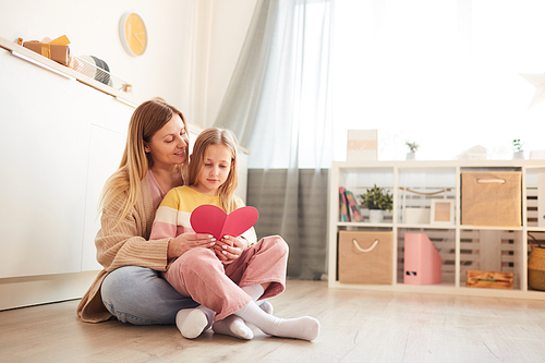 Full length portrait of mother and daughter holding present while sitting on floor in cozy childrens room interior, copy space