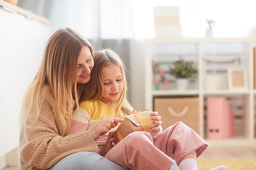 Warm-toned portrait of mother and daughter holding present while sitting on floor in cozy childrens room interior, copy space