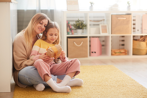 Full length portrait of mother and daughter holding present while sitting on floor in cozy childrens room interior, copy space