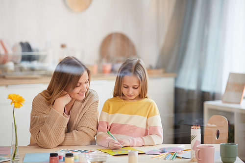 Warm-toned portrait of mature mother helping cute daughter with art and craft project in home interior, copy space