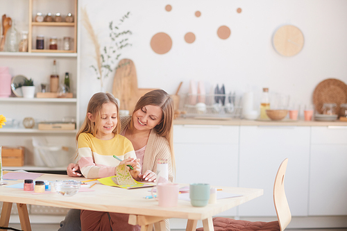 Warm-toned portrait of happy mother hugging little daughter while drawing pictures at wooden kitchen table, copy space