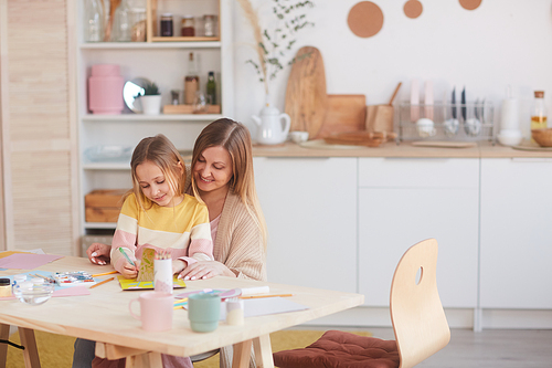 Warm-toned portrait of happy mother hugging little daughter while painting pictures at wooden table in kitchen, copy space