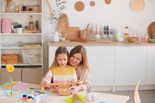 Warm-toned portrait of happy mother hugging daughter while opening presents at wooden kitchen table, copy space