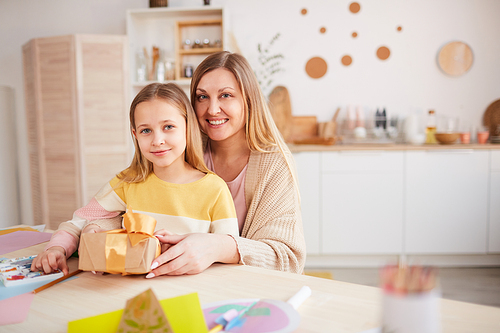 Warm-toned portrait of happy mother posing with daughter while opening presents at wooden kitchen table, copy space