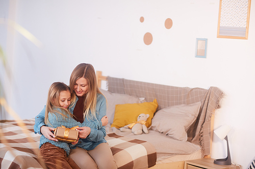 Wide angle portrait of happy mother hugging cute girl while opening present sitting on bed, copy space