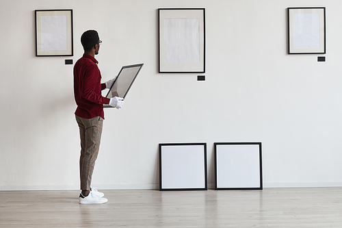 Full length portrait of African-American man planning art gallery or exhibition while setting up frames on white wall, copy space