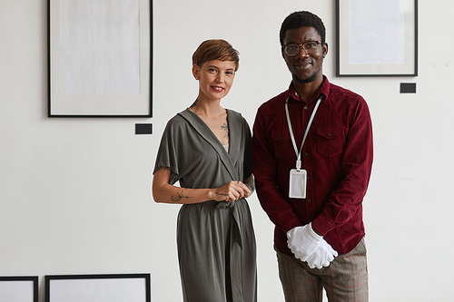 Portrait of female art gallery manager posing with African-American worker while standing against white wall and smiling at camera, copy space