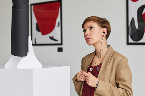 Portrait of contemporary young woman looking at sculptures and listening to audio guide at art gallery exhibition, copy space