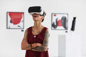 Waist up portrait of tattooed young woman wearing VR gear while enjoying immersive experience at modern art gallery exhibition, copy space