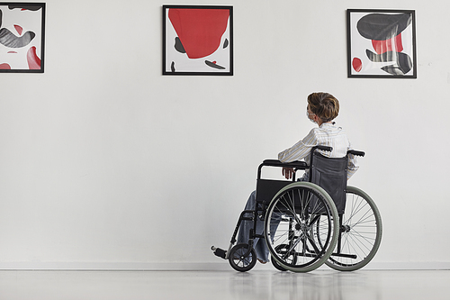 Wide angle portrait of young woman using wheelchair looking at paintings in modern art gallery, copy space