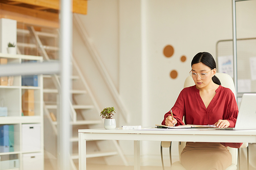 Portrait of successful Asian businesswoman writing at desk in modern white office interior, copy space