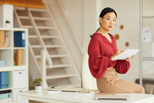 Portrait of elegant Asian businesswoman sitting on desk and  while working in modern white office, copy space