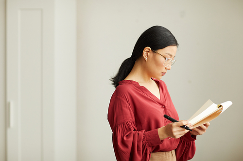 Side view portrait of successful Asian businesswoman wearing red blouse reading notes on clipboard while standing against white wall in office, copy space
