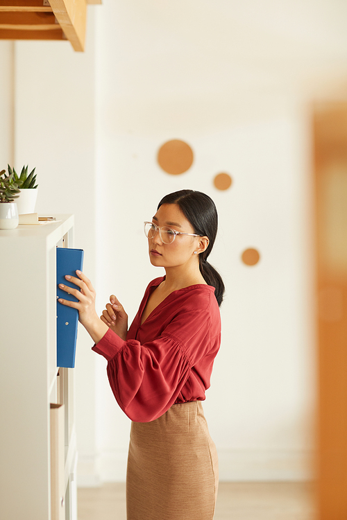 Side view portrait of elegant Asian businesswoman putting binder to bookshelf against white wall in office, copy space