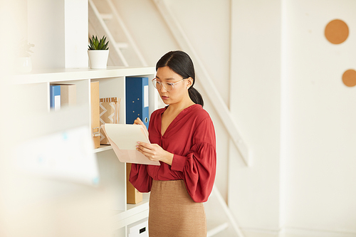 Portrait of elegant Asian businesswoman writing on clipboard while standing by bookshelves in office, copy space