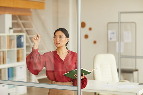 Waist up portrait of modern Asian businesswoman writing on glass wall while planning project in office, copy space