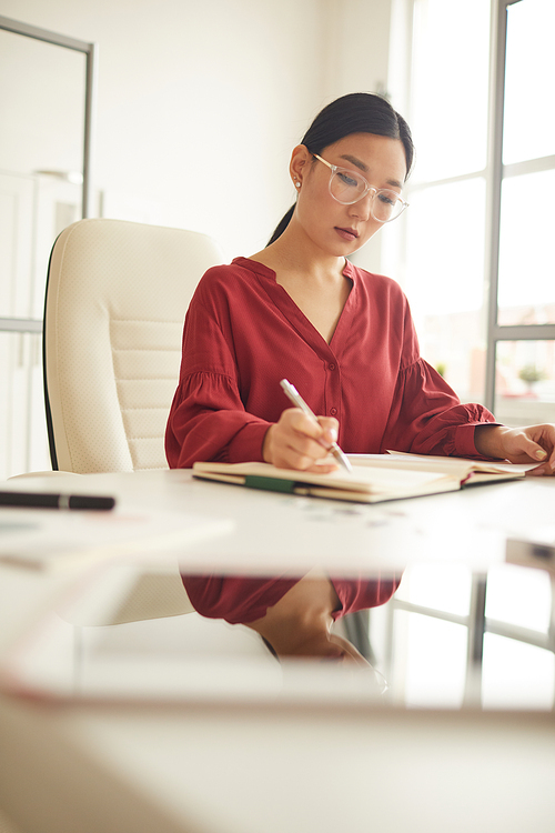Vertical portrait of elegant Asian woman reading documents while working at desk in modern white office, female boss concept, copy space