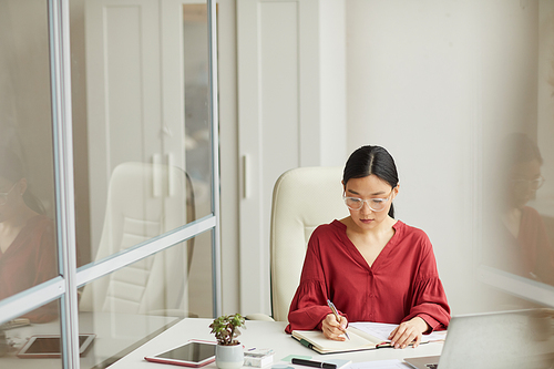Portrait of modern Asian businesswoman working at desk in white office sitting by glass wall with reflection, copy space