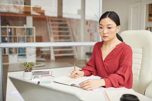 Portrait of modern Asian businesswoman wearing red blouse working at desk in office cubicle, copy space