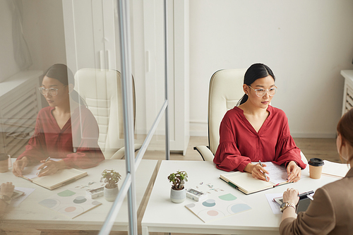 High angle view at modern Asian businesswoman talking to client while working at desk in white office cubicle, copy space