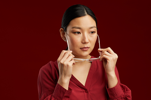 Head and shoulders portrait of elegant Asian woman putting on glasses while standing against maroon background in studio, copy space