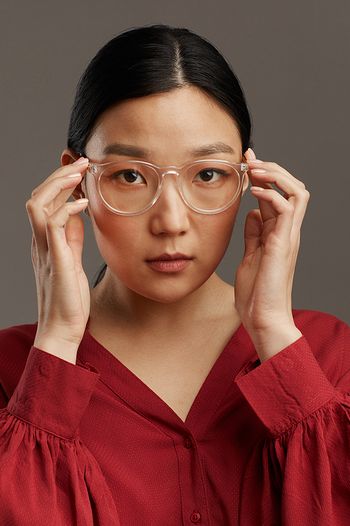 Head and shoulders portrait of elegant Asian woman putting on glasses while standing against grey background in studio