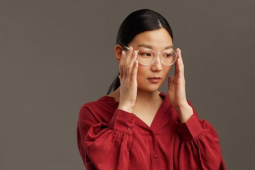 Head and shoulders portrait of elegant Asian woman putting on glasses while standing against grey background in studio, copy space
